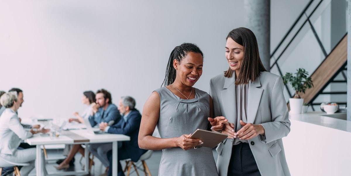 two women in office looking at tablet, group of people sitting at table in thebackground